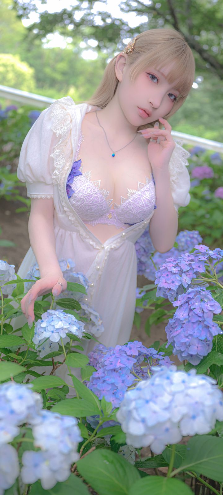 A blonde woman in a white lace dress poses among blooming blue and purple hydrangeas in a garden.