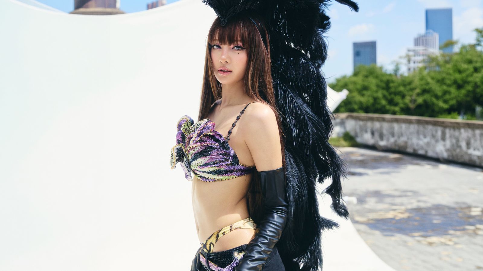 A glamorous woman in a sequined carnival costume with large black feather wings poses on a rooftop with a city skyline behind.