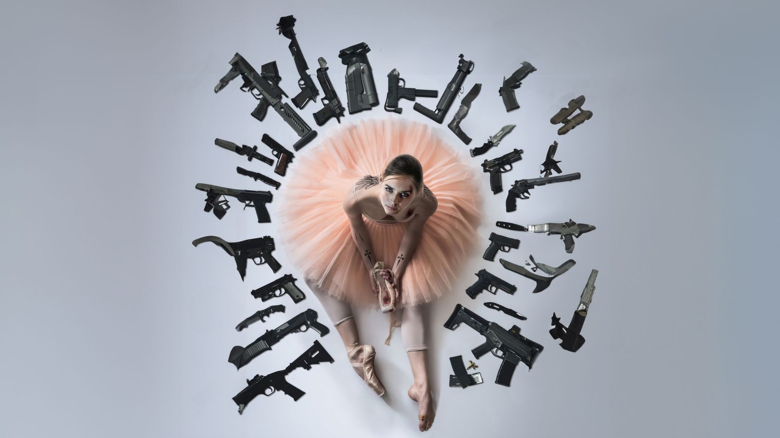 A ballerina in a peach tutu surrounded by a circular arrangement of guns and weapons on a light gray background.
