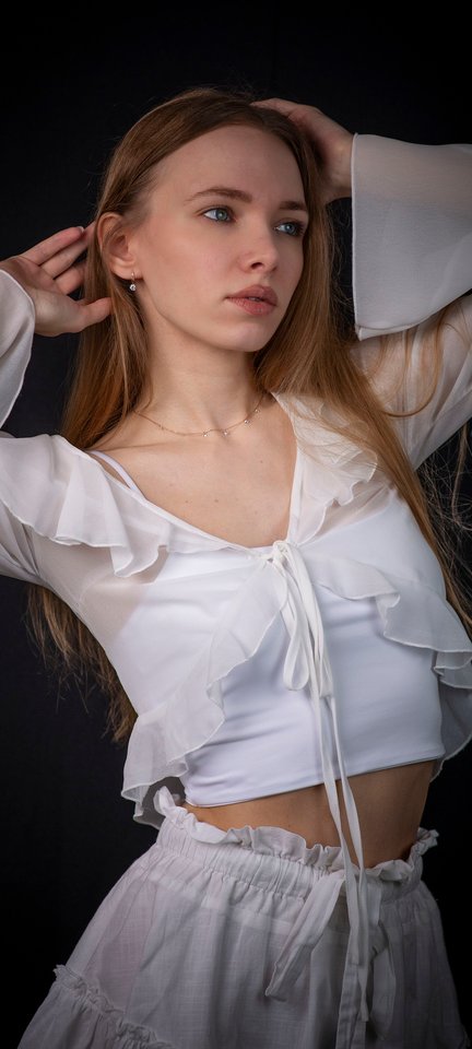 A young woman with long light brown hair in a white ruffled outfit posing against a dark background.