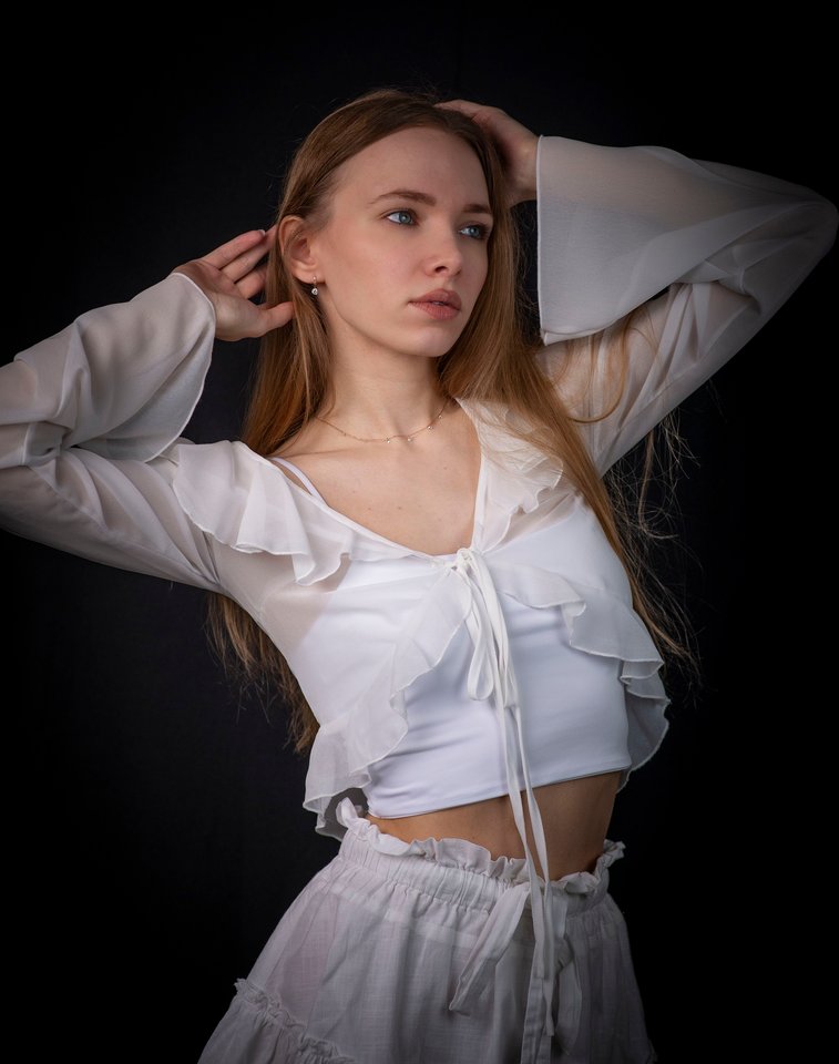 A young woman with long light brown hair in a white ruffled outfit posing against a dark background.